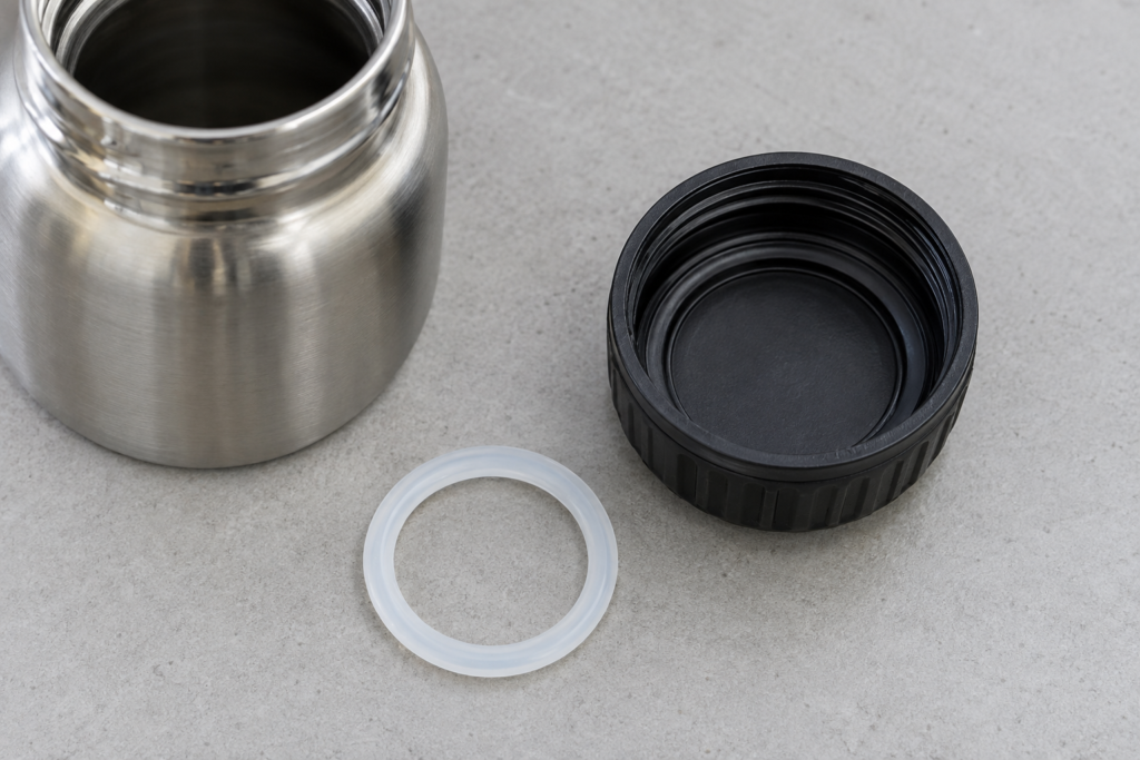 Gloved hand holding a bottle cap with its sealing ring visible beside a threaded stainless steel bottle neck.